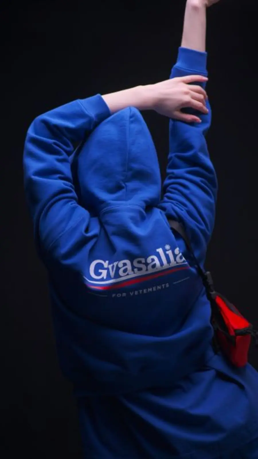 A person in a blue hoodie poses with arms raised against a dark background, displaying modern streetwear fashion.