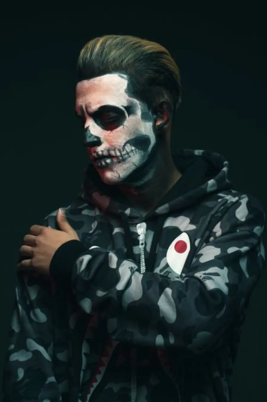 Artistic portrait of a man with skull face paint in a studio setting, wearing a camouflage hoodie.