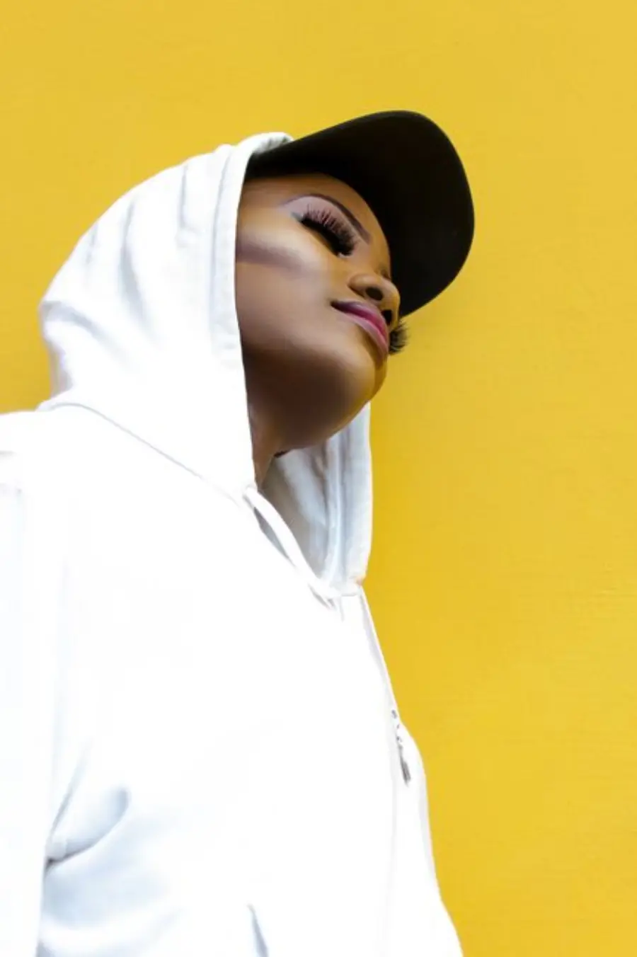 Confident woman posing in a white hoodie and cap against a yellow background.