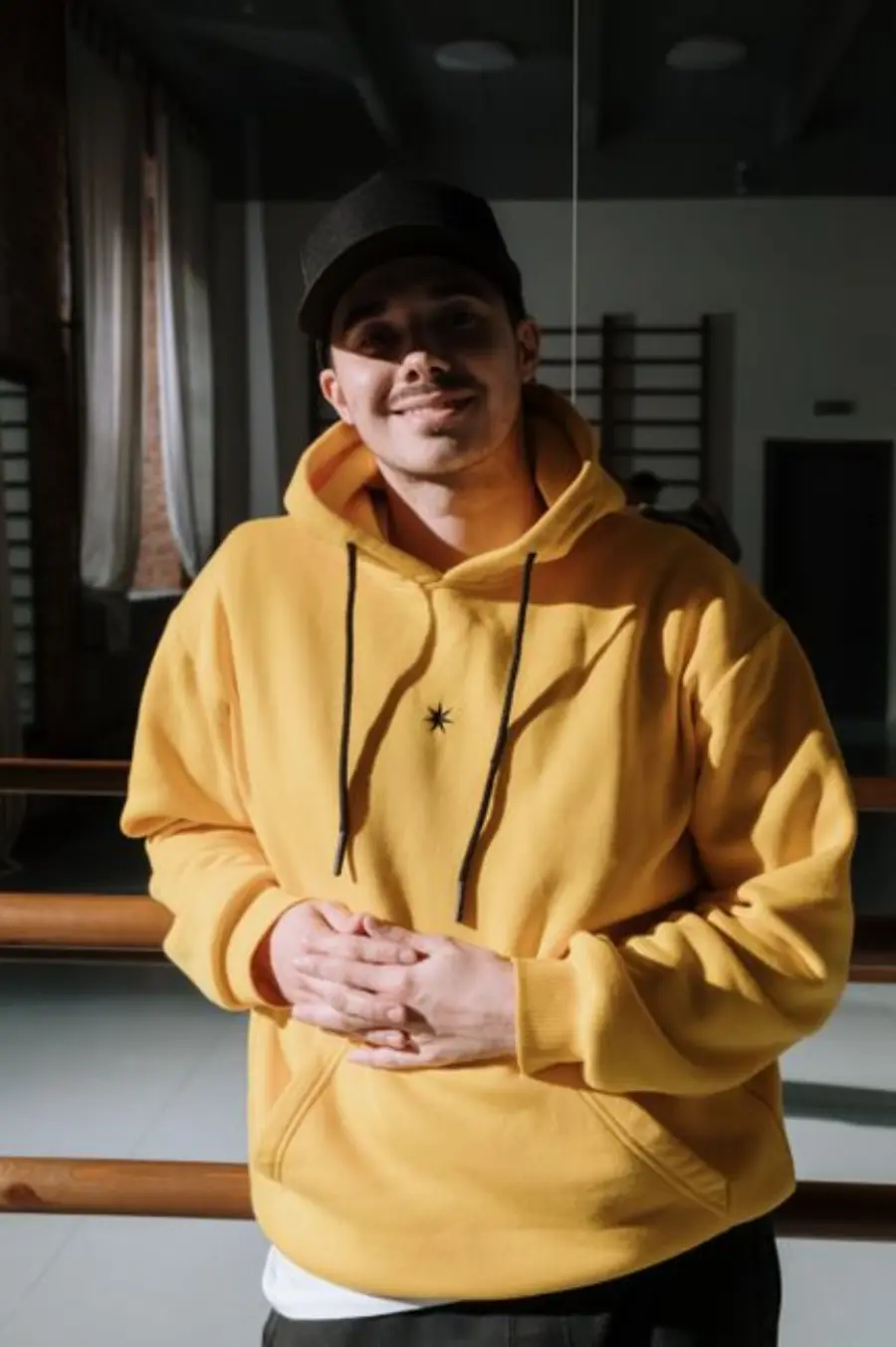 Smiling man in a yellow hoodie indoors, standing confidently in a dance studio.